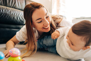 Happy young mother and adorable content baby wearing casual clothes lying on floor and playing together in living room - ADSF26807