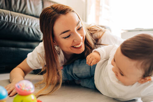 Glückliche junge Mutter und entzückendes zufriedenes Baby in Freizeitkleidung, das auf dem Boden liegt und zusammen im Wohnzimmer spielt - ADSF26807