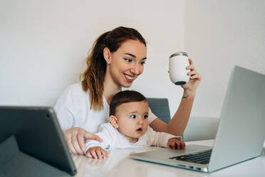 Glückliche junge Mutter und neugieriges Baby in weißen Hemden sehen sich ein lustiges Video auf einem Netbook an, während sie zusammen am Schreibtisch in einem hellen Raum sitzen - ADSF26808