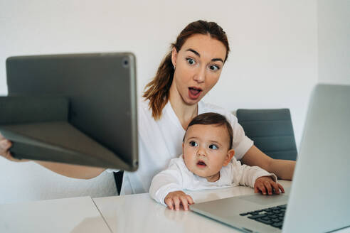 Erstaunte junge Mutter und süßes Baby sitzen am Schreibtisch und Surfen Netbook und Tablet zusammen in hellen Raum - ADSF26809