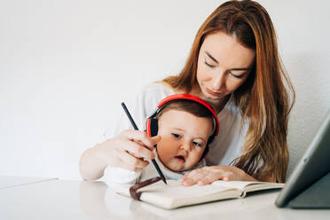 Positive junge Mutter mit Bleistift, die einem liebenswerten, fleißigen Baby mit Kopfhörern beibringt, in ein Tagebuch zu schreiben, während sie zusammen am Schreibtisch in einem hellen Raum sitzen - ADSF26811