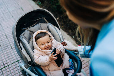 Von oben niedlich lustig Baby mit Smartphone tragen warme Kleidung sitzen im Kinderwagen und Blick auf unscharfe Mutter auf Frühling Straße - ADSF26819