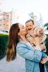 Cheerful young mother in casual clothes carrying and gently kissing adorable joyful baby while standing on city sidewalk on sunny spring day - ADSF26820