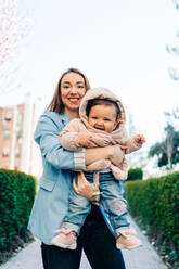 Fröhliche junge Mutter in Freizeitkleidung schaut in die Kamera und trägt ein entzückendes, fröhliches Baby, während sie an einem sonnigen Frühlingstag auf dem Bürgersteig der Stadt steht - ADSF26821