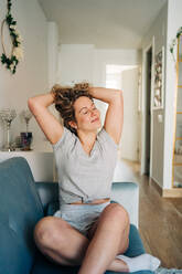 Positive attraktive Frau in Shorts sitzt auf einer gemütlichen Couch im Wohnzimmer und streckt die Arme mit geschlossenen Augen nach oben - ADSF26822