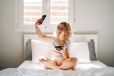 Glad young female in domestic clothes taking selfie pictures of yummy breakfast in bowl while sitting with legs crossed on cozy bed in morning - ADSF26829