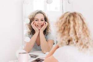 Glücklich lächelnde attraktive Frau im Freizeithemd, die ihr Gesicht berührt und ihr Spiegelbild betrachtet, während sie am Frisiertisch im Schlafzimmer sitzt - ADSF26831