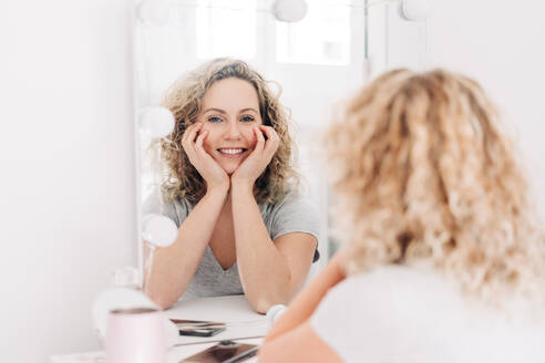 Happy smiling attractive female in casual shirt touching face and looking at reflection in mirror while sitting at vanity table in bedroom - ADSF26831