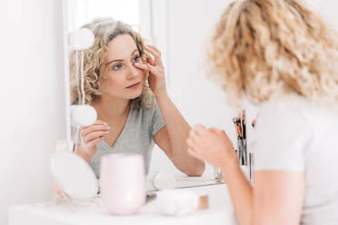 Content young female in casual shirt applying moisturizing cream on face and looking in mirror while sitting at vanity table in light bedroom - ADSF26833