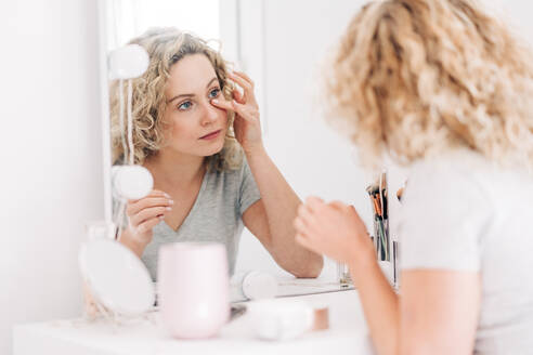 Content young female in casual shirt applying moisturizing cream on face and looking in mirror while sitting at vanity table in light bedroom - ADSF26833