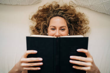 From above happy young female with curly hair lying on bed reading book during weekend at home - ADSF26838