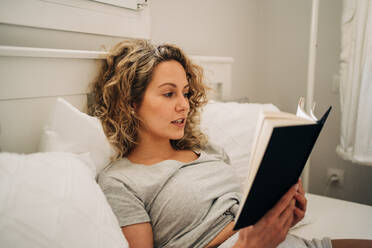 Young female with curly hair lying on bed reading book during weekend at home - ADSF26839