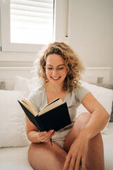 Positive young female with curly blond hair in panties and eyeglasses smiling while sitting on cozy bed and reading interesting book - ADSF26840