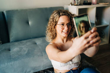 From above of self assured young female millennial with curly blond hair in leather bra an jeans sitting on floor near sofa and taking selfie on smartphone - ADSF26842