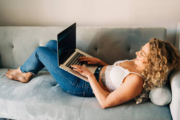 Seitenansicht einer jungen selbständigen Frau in Freizeitkleidung und mit Brille, die auf einem bequemen Sofa liegt und aus der Ferne an einem Laptop arbeitet - ADSF26843