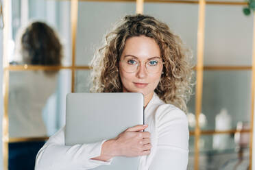 Self assured young female entrepreneur with curly blond hair in white blouse and eyeglasses looking at camera while standing in modern workspace with laptop in hands - ADSF26845