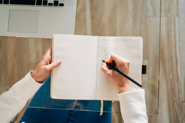 From above of crop anonymous female student with curly hair in casual clothes sitting at glass table and taking notes in copybook while preparing for exams using laptop at home - ADSF26848