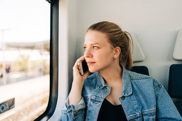 Junge, lässige Frau in Jeansjacke, die telefoniert und dabei aus dem Fenster eines Zuges schaut - ADSF26851