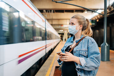 Young female in casual clothes and protective mask standing on railway station and looking away while waiting for train - ADSF26853