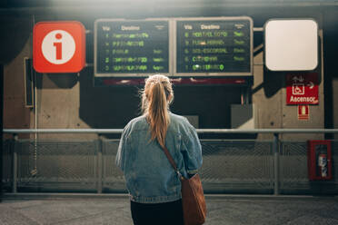 Rückenansicht einer Frau in Jeansjacke und mit Tasche, die auf einem Bahnhof stehend den Fahrplan der Züge an Bord betrachtet - ADSF26854