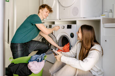 Cheerful young guy in casual outfit loading clothes in washing machine and smiling while communicating with positive girlfriend using smartphone in modern laundry room - ADSF26856