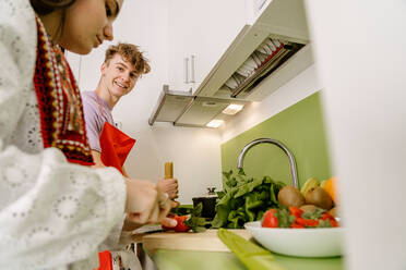 Low angle of crop ethnic female in stylish outfit cutting fresh strawberry on chopping board while cooking in kitchen with cheerful boyfriend - ADSF26858