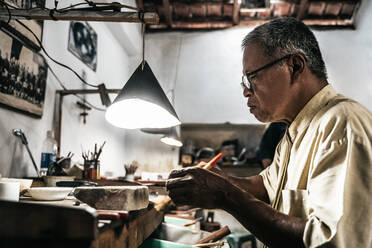 Side view of mature ethnic man working at weathered desk in craft workshop - ADSF26866