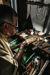 Von oben konzentrierter ethnischer älterer männlicher Juwelier in Freizeitkleidung und mit Brille, der Instrumente schärft, während er an einem Holztisch in einer traditionellen Werkstatt arbeitet - ADSF26869