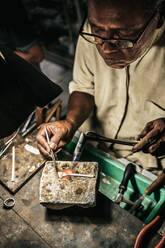 Reifer ethnischer Mann mit brennendem Brenner, der ein Metallstück erhitzt, das am Schreibtisch in einer Handwerkswerkstatt bearbeitet wird - ADSF26870