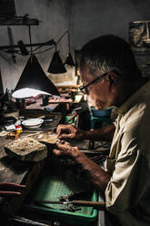Seitenansicht eines reifen ethnischen Mannes, der an einem verwitterten Schreibtisch in einem Handwerksbetrieb arbeitet - ADSF26871