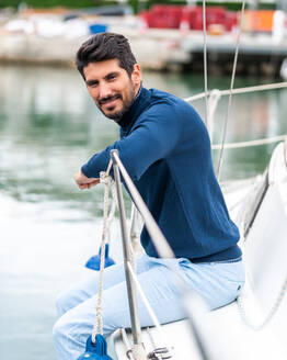 Seitenansicht eines zufriedenen ethnischen Mannes mit Bart, der in die Kamera schaut, während er auf einer Yacht auf einem See im Hafen sitzt - ADSF26874