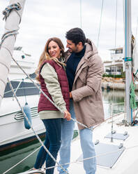 Cheerful multiethnic couple in outerwear embracing and talking on yacht on river in urban harbor - ADSF26875