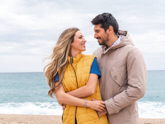 Smiling woman with cheerful ethnic male beloved looking at each other on ocean beach in daylight - ADSF26884