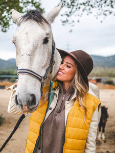 Glückliche junge Reiterin in trendigem Outfit und Hut lächelnd, während sie ein liebenswertes reinrassiges graues Pferd auf dem Lande streichelt - ADSF26890