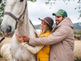 Seitenansicht von romantischen jungen ethnischen Paar in stilvoller Kleidung umarmt und streichelt gehorsam grauen Pferd auf bewölkten Tag in Ranch - ADSF26891