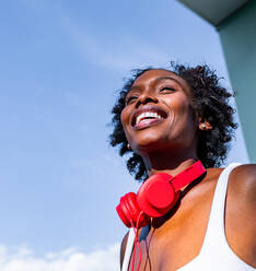 Joyful African American female in white top and red shorts with headphones on neck leaning on white wall and laughing on sunny day - ADSF26908