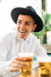 Cheerful stylish tomboy with tattoos wearing hat and white shirt drinking beer from glass in cafe - ADSF26915