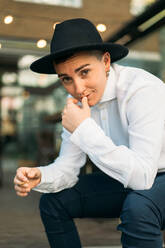 Smiling masculine female in stylish hat and white shirt smoking and looking at camera - ADSF26918