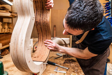 Adult male artisan making classical musical instrument with wooden pieces and clamp in workshop - ADSF26928