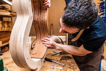 Erwachsener männlicher Kunsthandwerker, der ein klassisches Musikinstrument mit Holzteilen und einer Klemme in einer Werkstatt herstellt - ADSF26928