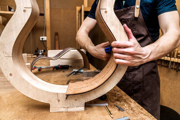 Abgeschnittener, nicht erkennbarer erwachsener männlicher Handwerker, der ein klassisches Musikinstrument mit Holzteilen und einer Klemme in einer Werkstatt herstellt - ADSF26929