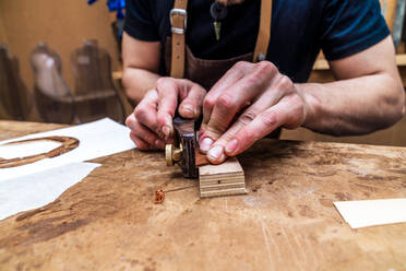 Anonymer, unrasierter männlicher Handwerker, der einen Holzklotz mit einem Handwerkzeug an einem Tisch in einem Arbeitsraum glättet - ADSF26930