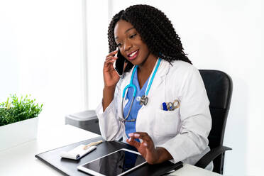 Positiver afroamerikanischer Arzt, der ein Gespräch über ein Mobiltelefon führt, während er in einem medizinischen Krankenhaus an einem Tisch mit einem Tablet arbeitet - ADSF26935