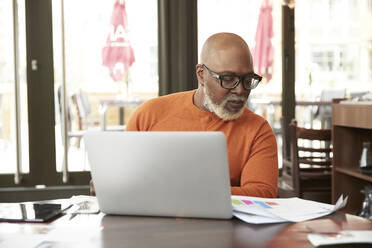 Glatzköpfiger Geschäftsmann, der in einem Restaurant an einem Laptop arbeitet und Dokumente betrachtet - PMF02030