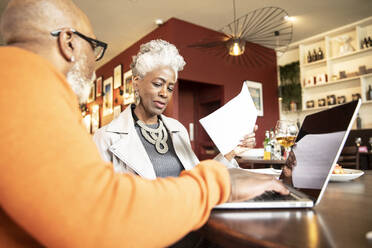 Ältere Frau diskutiert mit einem männlichen Kollegen, der im Restaurant am Laptop arbeitet - PMF02043