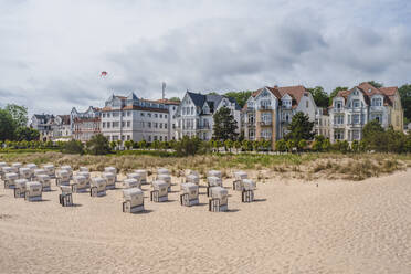 Germany, Mecklenburg-Western Pomerania, Heringsdorf, Hooded beach chairs on sandy beach with residential buildings in background - KEBF01999