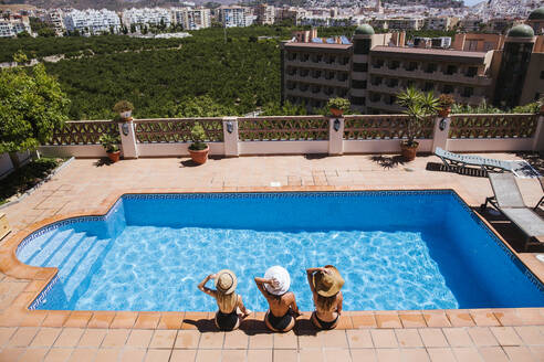 Female friends wearing sun hats sitting at poolside on sunny day - LJF02216