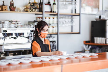 Waitress wearing protective face mask arranging cup on saucer at cafeteria counter - LJF02222