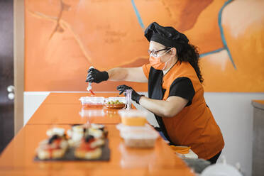 Köchin mit Gesichtsschutzmaske beim Verpacken von Lebensmitteln in einem Container in einer Cafeteria - LJF02238