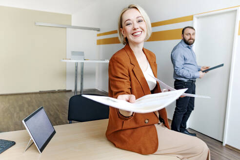Fröhliche blonde Geschäftsfrau, die Dokumente verteilt, während sie auf dem Schreibtisch im Büro sitzt - OGF01051